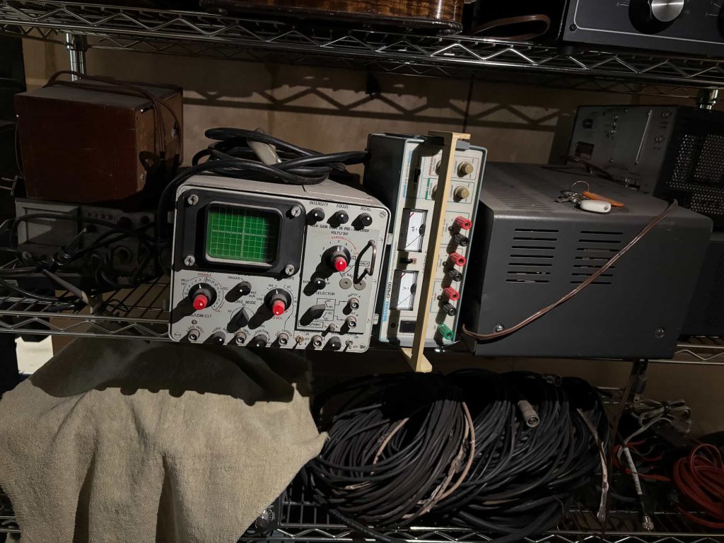 Vintage electronic equipment on metal shelves, featuring an oscilloscope with a green screen and red knobs. Coiled cables and cloth lie below, evoking a retro workshop ambiance.