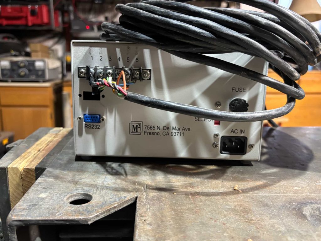 Rear panel of an M2 electronic device featuring a six-position screw terminal block with wires connected to terminals 1 through 4, a blue RS232 port, a fuse holder, a red selector switch, and an IEC C14 AC power input.