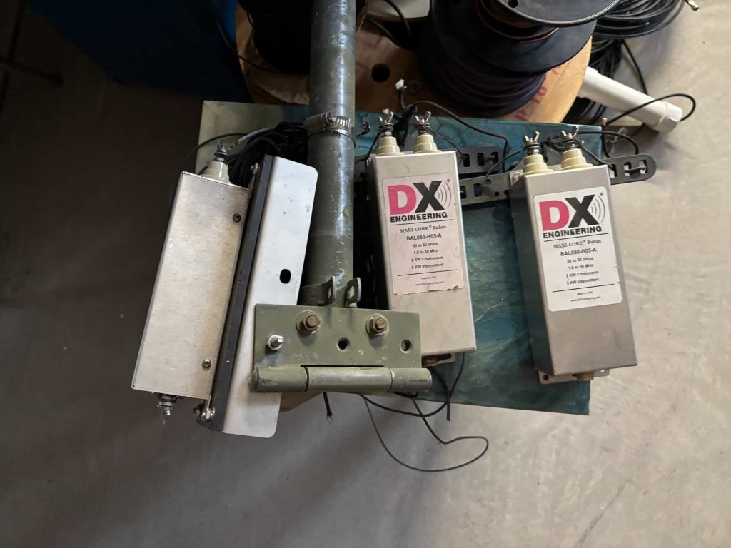 Three DX Engineering Maxi-Core BAL050-H05-A baluns mounted to a central grey metal pole. Two of the silver rectangular enclosures display labels with specifications: 50 to 50 ohms, 1.8 to 30 MHz, 2 kW continuous, and 5 kW intermittent power ratings. The units feature top-mounted ceramic insulators with wing-nut terminals. A green heavy-duty hinged mounting bracket is attached to the central pole, which is secured with a hose clamp.