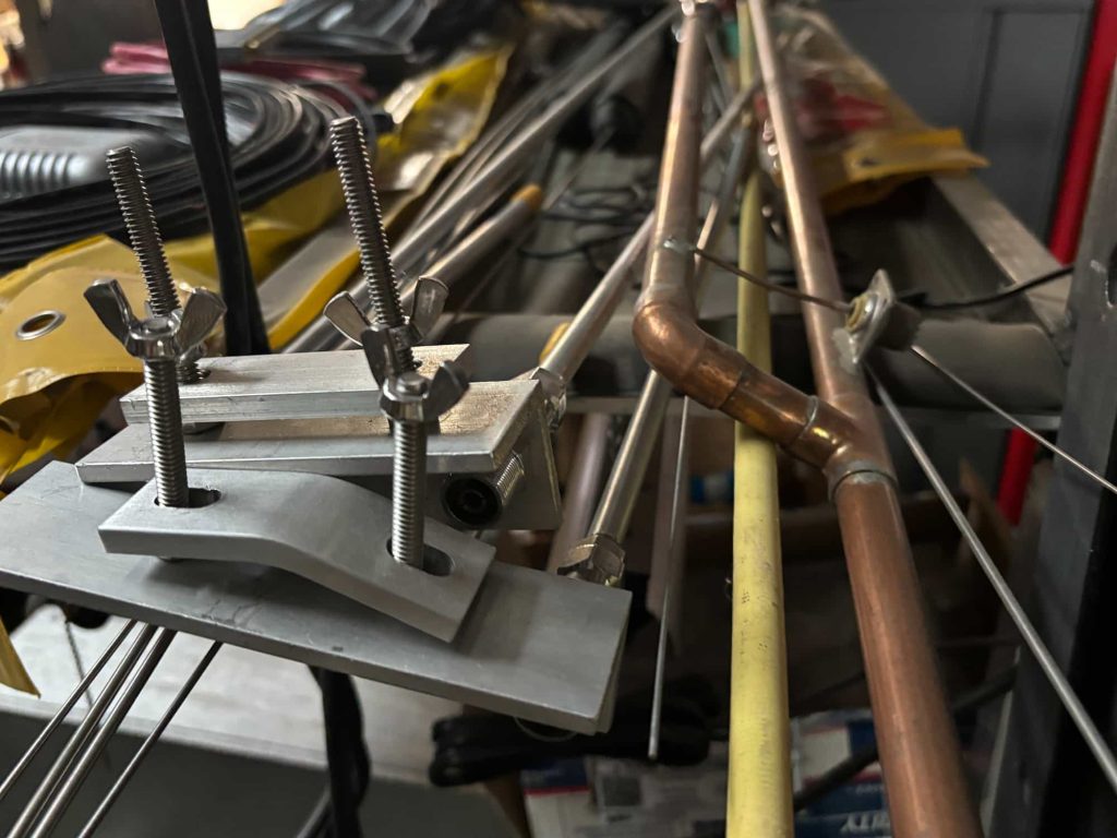 Close-up of a cluttered workshop containing various metal components. In the foreground, two vertical threaded studs with wing nuts secure a pair of aluminum mounting plates. To the right, a copper pipe assembly with an elbow joint runs parallel to a yellow flexible conduit. The background is filled with various metal rods, tubes, and tools.