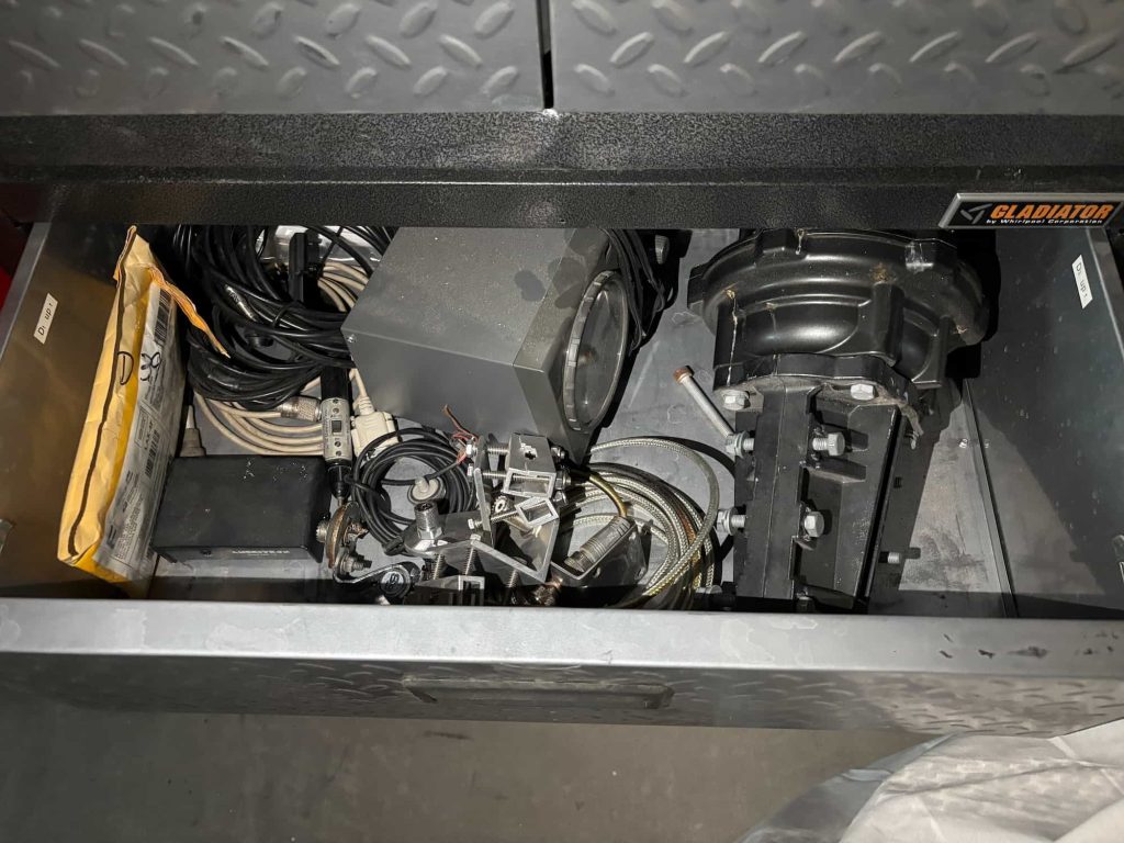 A metal drawer labeled "Gladiator" containing various electrical cables, a black device, a yellow envelope, and mechanical parts.