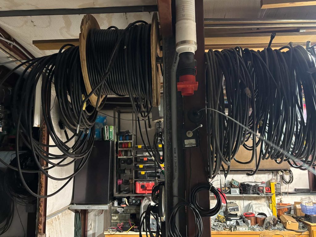 Multiple coils and a wooden spool of heavy-duty black cable hang from overhead supports in a cluttered workshop. A central vertical metal post features a white PVC conduit, a red and black industrial power connector, and a small control box with a toggle switch. The background shows workbenches and shelves filled with various tools, electronic components, and parts bins.