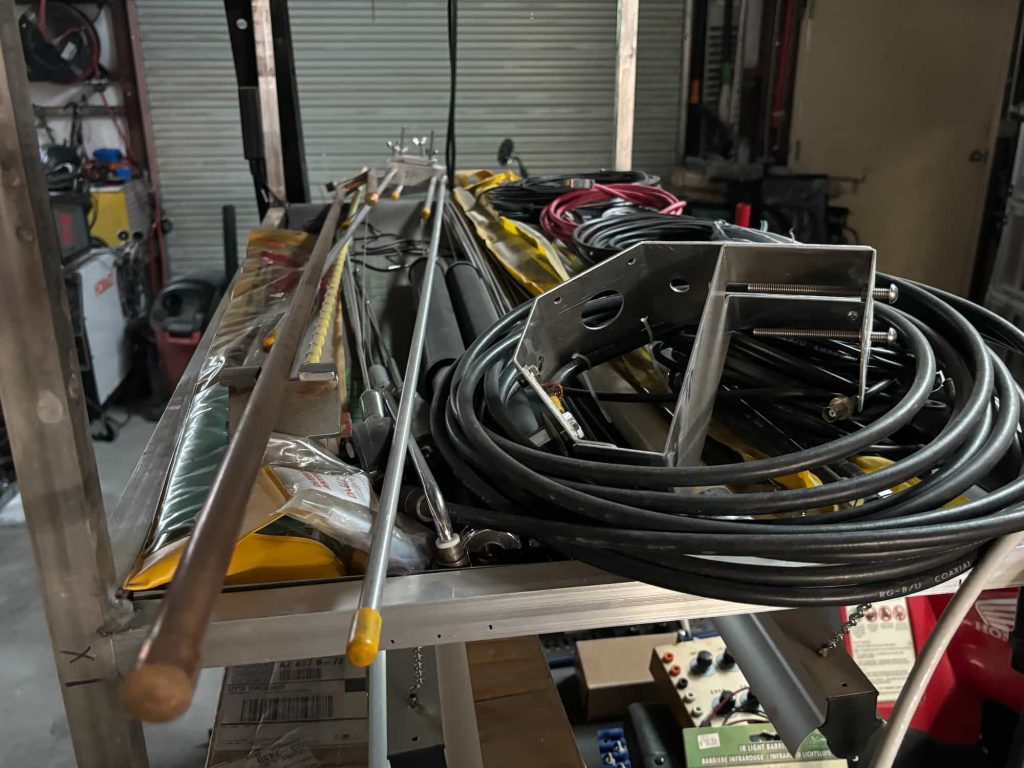 A metal equipment cart holding a large coil of black RG-8/U coaxial cable, a stainless steel antenna mounting bracket, several thin whip antenna rods with yellow tips, and various packaged antenna components in a workshop.