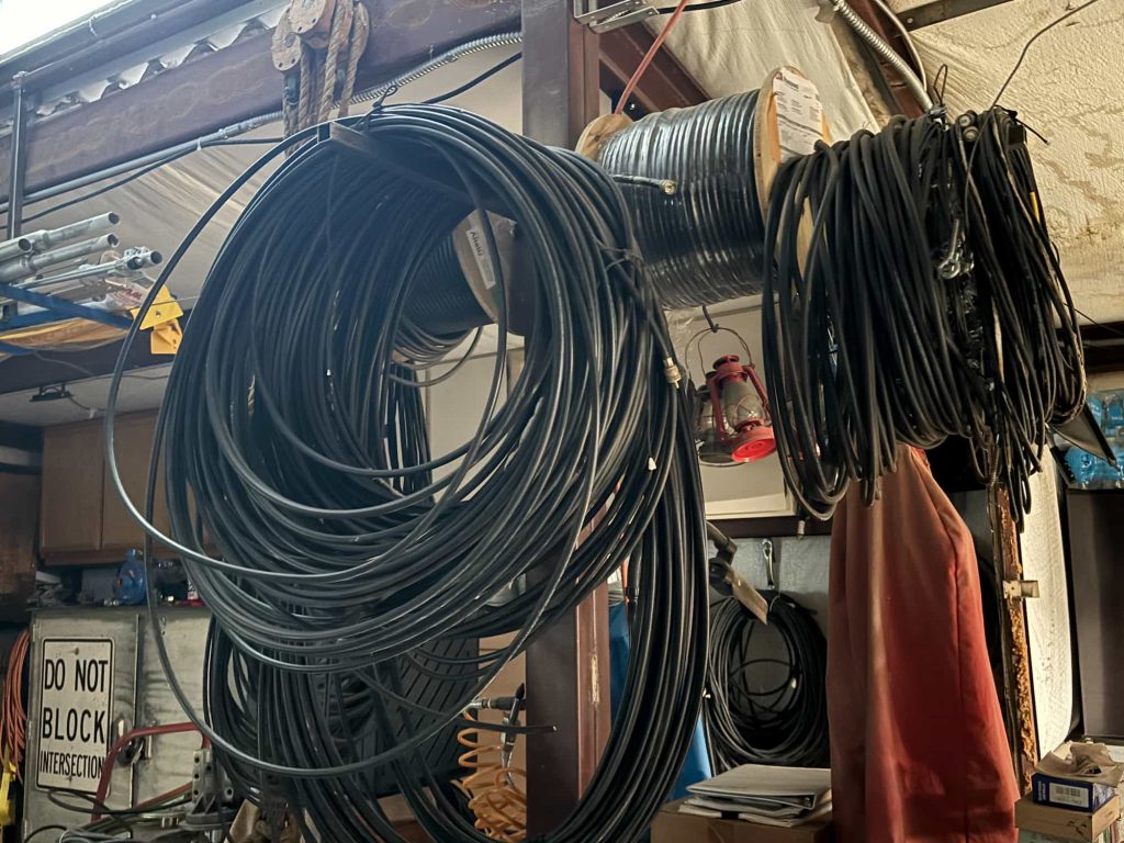 Large coils and a spool of thick black cable hanging from horizontal metal support beams in an industrial workshop. A "DO NOT BLOCK INTERSECTION" sign and a red lantern are visible in the background among various tools and equipment.