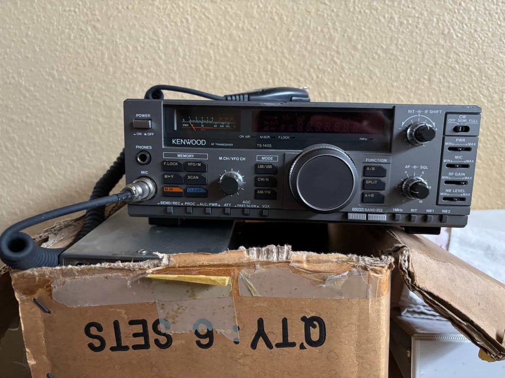 A Kenwood radio transceiver sits on top of a worn cardboard box labeled "TV 6 SETS," with a coiled microphone attached on the left.