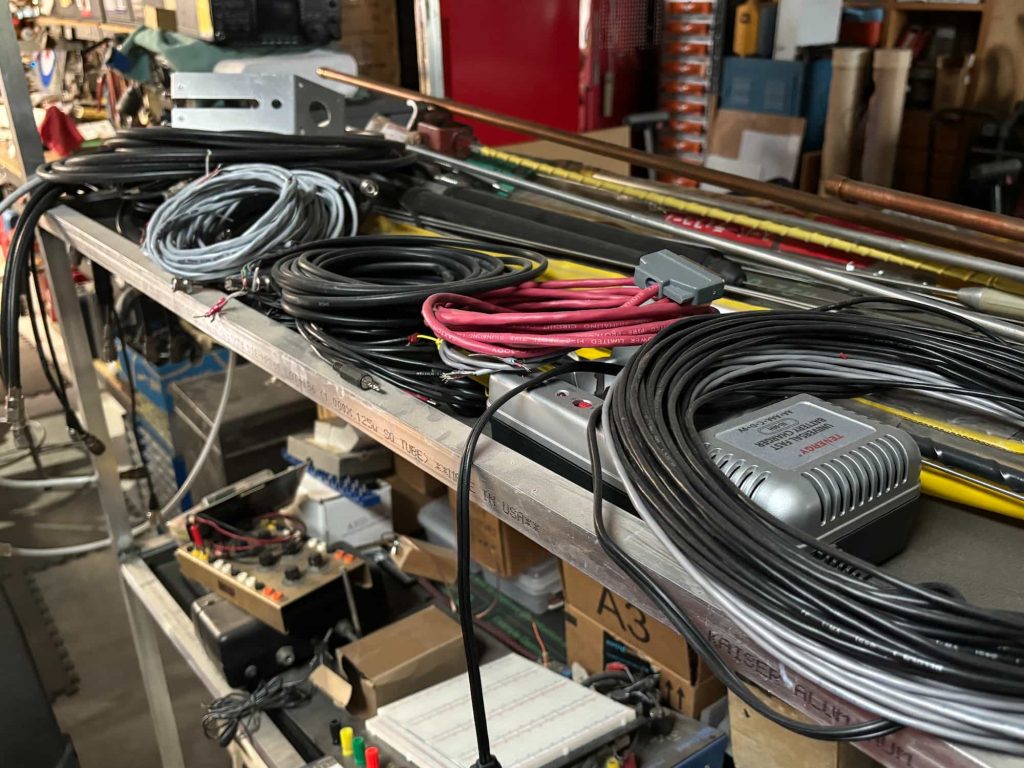 A metal workbench shelf holds several coils of black coaxial cable, gray wire, and red multi-conductor cable with a gray connector. A Tenergy universal fast battery charger and an aluminum mounting bracket are positioned among the cables. Long copper pipes and yellow rods rest horizontally behind the wire coils. A lower shelf contains an electronic breadboard and various components.