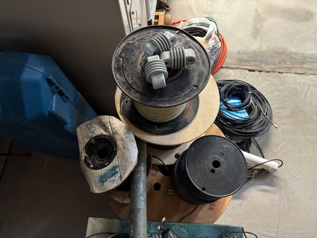 A top-down view of multiple spools on a concrete floor. A large black reel holding light-colored wire has three grey, ribbed ceramic insulators sitting on top. Adjacent is a smaller spool with a torn white cardboard cover labeled "Antenna Rope" and another empty black plastic reel. Coiled black and orange cables, a blue tool case, and a galvanized steel pipe are visible in the background.