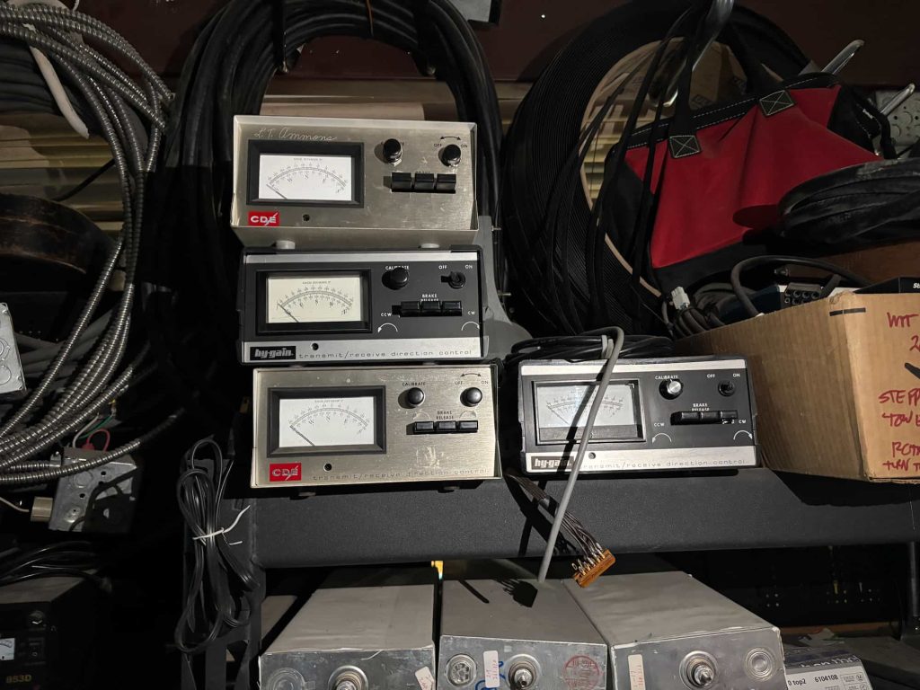 Stacked vintage analog amplifiers with dial indicators and buttons, surrounded by coiled cables, a red bag, and a cardboard box on a dimly lit shelf.