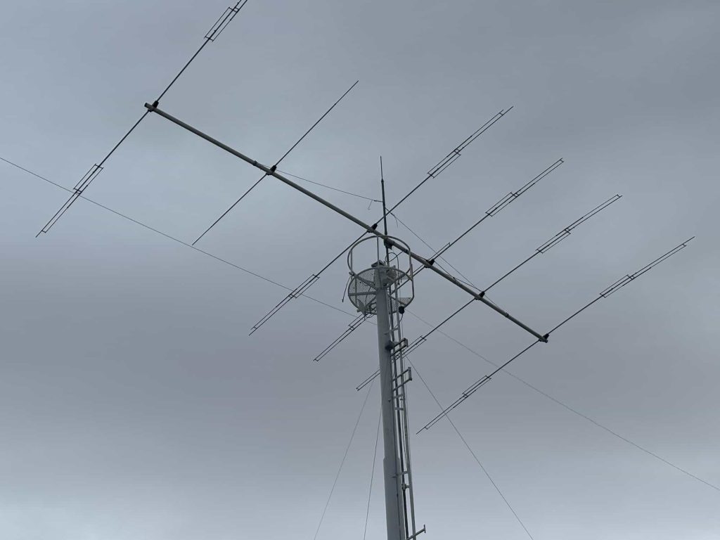 A multi-element Yagi or log-periodic antenna array mounted atop a tall vertical metal mast. The mast is supported by guy wires and features additional smaller antennas and a small circular platform near the top. The structure is situated behind a building's roofline against an overcast, grey sky.
