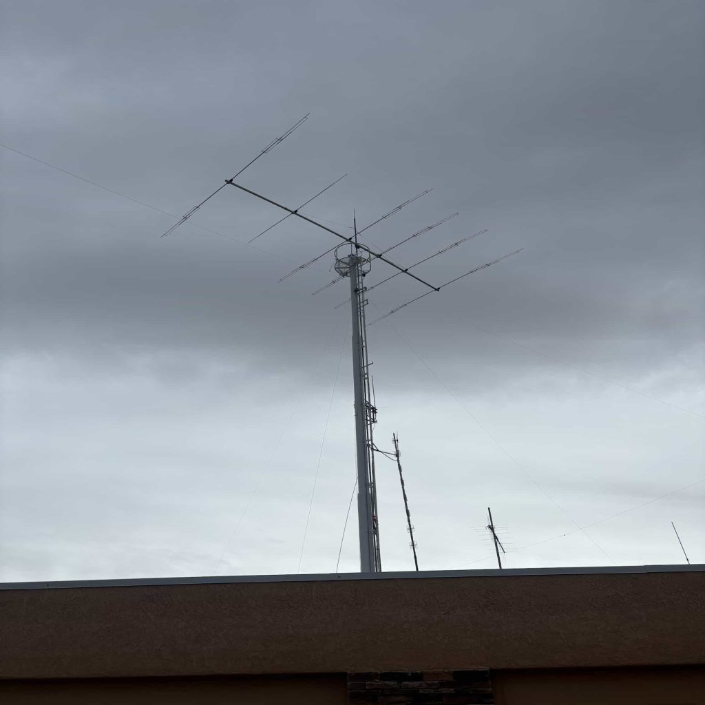 A multi-element Yagi or log-periodic antenna array mounted atop a tall vertical metal mast. The mast is supported by guy wires and features additional smaller antennas and a small circular platform near the top. The structure is situated behind a building's roofline against an overcast, grey sky.
