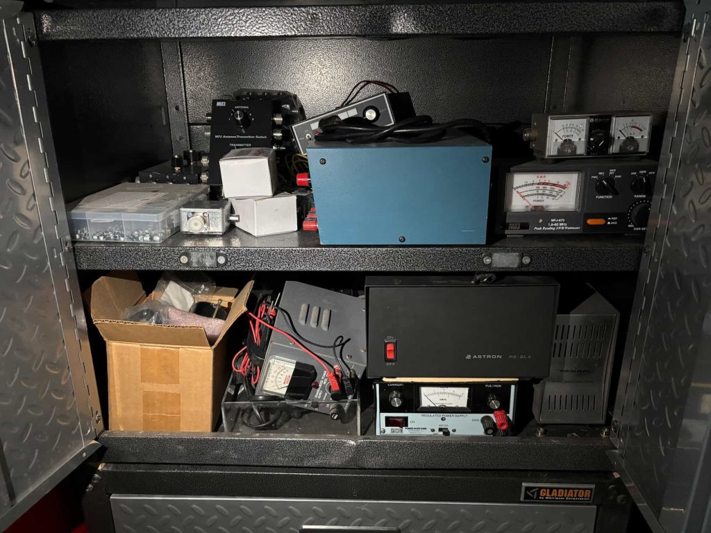 Metal cabinet filled with electronic equipment. Shelves hold meters, boxes, wiring, and a blue device, conveying a sense of organized chaos.