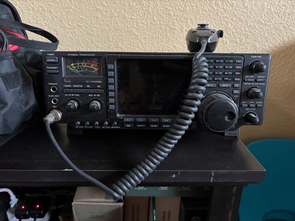 A radio transceiver with a coiled microphone cord and various buttons sits on a dark wooden table against a beige wall, conveying a vintage vibe.