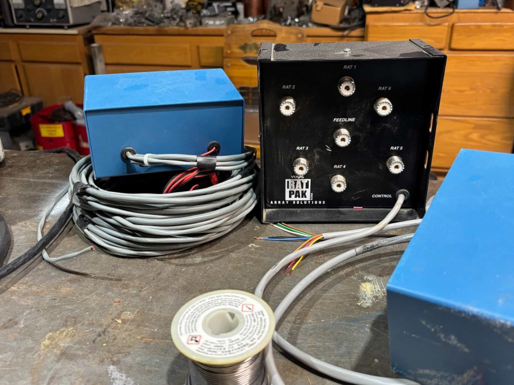 An Array Solutions Rat Pak antenna switch featuring six SO-239 connectors labeled RAT 1 through 6 around a central Feedline port. A blue control box sits atop a large coil of grey multi-conductor cable. Stripped control wires and a spool of solder are in the foreground on a metal workbench.