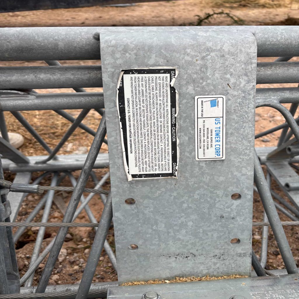 Galvanized steel lattice tower section with an attached flat metal bracket. The bracket holds two labels: a safety warning label titled "DANGER / WARNING - CAUTION" and a manufacturer's identification label for "US TOWER CORP." containing contact information and a "Made in the USA" logo.