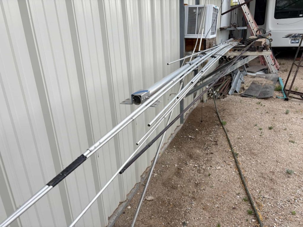 Multiple aluminum antenna elements and tubes leaning against a white corrugated metal wall. A primary boom section features a square mounting bracket with a recessed electrical connector.